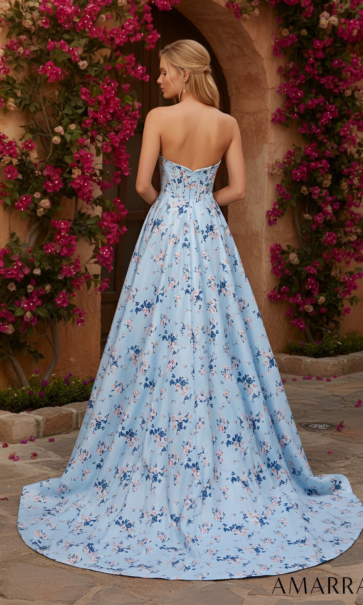 A woman stands outdoors facing away from the camera, wearing the Long Prom Dress: Amarra 89614—a strapless, floor-length blue gown. The background features pink flowers and greenery.